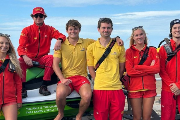 Six RNLI lifeguards in red and yellow uniforms posing with a rescue watercraft on the promenade at Aberavon Beach