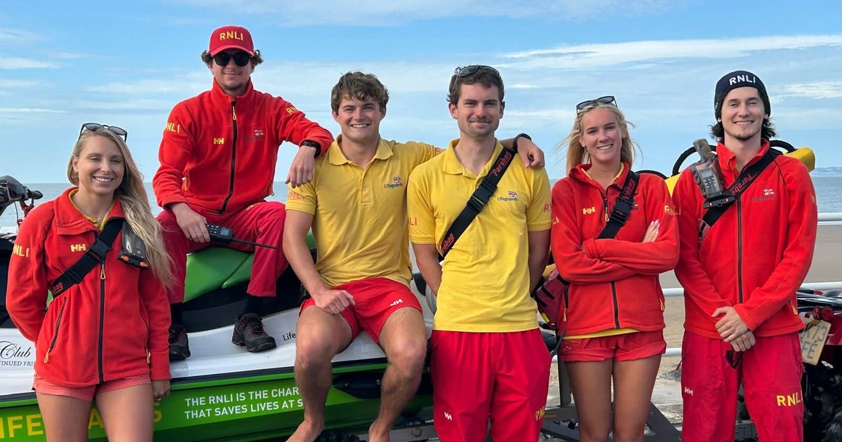 Six RNLI lifeguards in red and yellow uniforms posing with a rescue watercraft on the promenade at Aberavon Beach