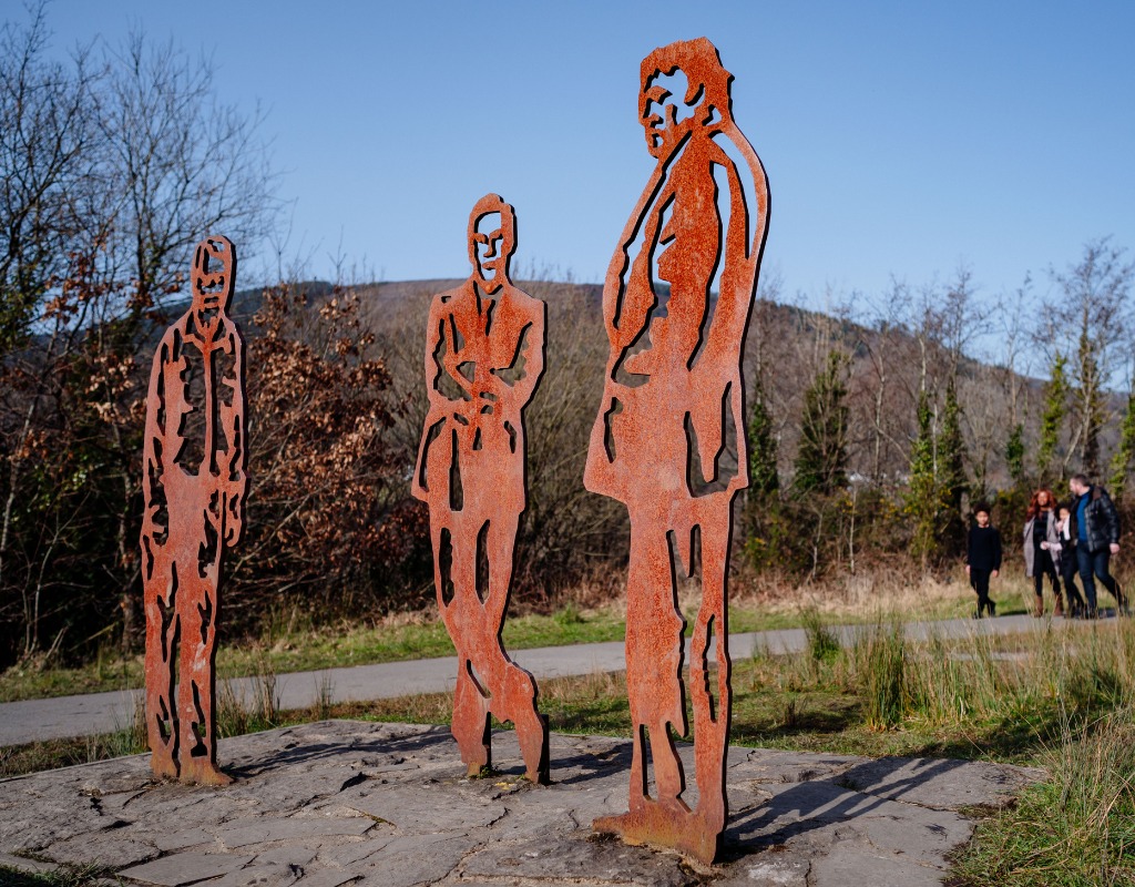 Three rusted steel cut-out figures of men standing on a stone base beside a path, with walkers and a hillside behind