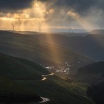 Shafts of sunlight over the Afan Valley, with a winding road, a small village and wind turbines on the ridge