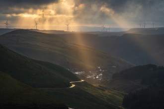 Shafts of sunlight over the Afan Valley, with a winding road, a small village and wind turbines on the ridge