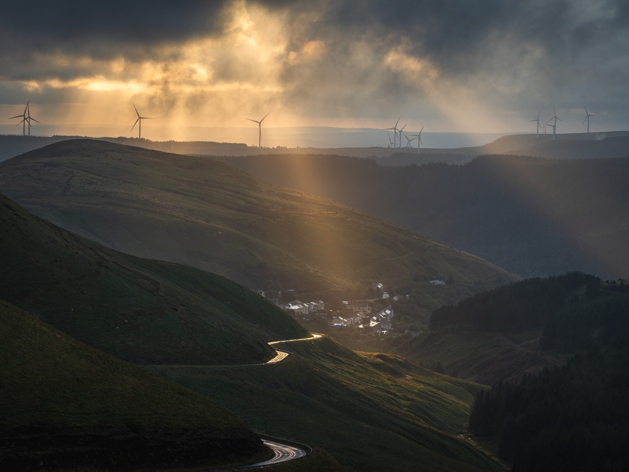 Shafts of sunlight over the Afan Valley, with a winding road, a small village and wind turbines on the ridge