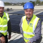 Two men in hi-vis vests and hard hats kneeling on a factory roof covered in black solar panels, with trees and hills behind