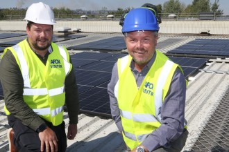 Two men in hi-vis vests and hard hats kneeling on a factory roof covered in black solar panels, with trees and hills behind