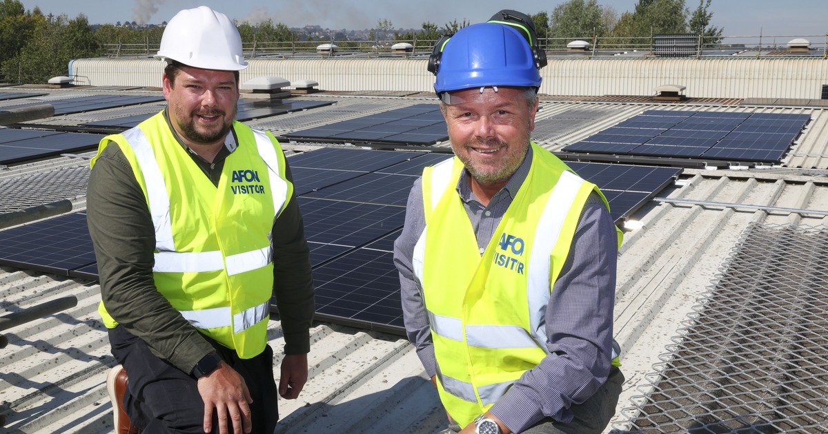 Two men in hi-vis vests and hard hats kneeling on a factory roof covered in black solar panels, with trees and hills behind