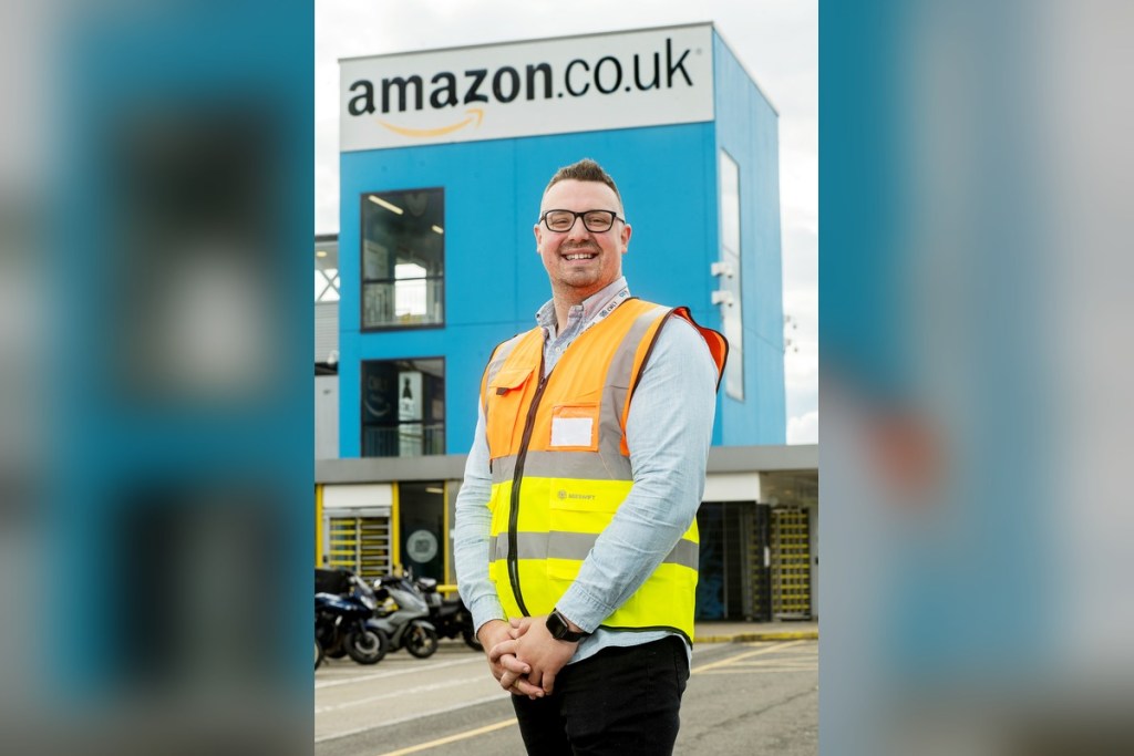 Chris Bailey smiling in a hi-vis vest outside the blue Amazon Swansea building beneath the amazon.co.uk sign
