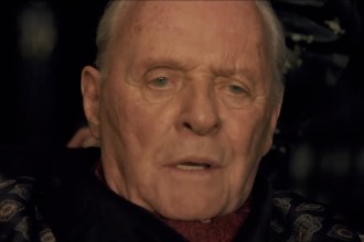 Close-up of Anthony Hopkins in a dark patterned robe and red cravat, speaking in dim light