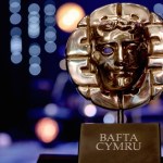 A gold BAFTA Cymru mask trophy on a black plinth against a dark stage with blue and orange lights out of focus behind it