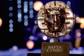 A gold BAFTA Cymru mask trophy on a black plinth against a dark stage with blue and orange lights out of focus behind it