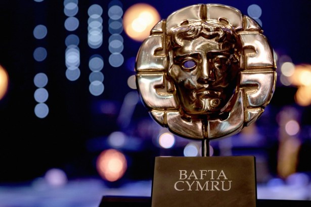 A gold BAFTA Cymru mask trophy on a black plinth against a dark stage with blue and orange lights out of focus behind it