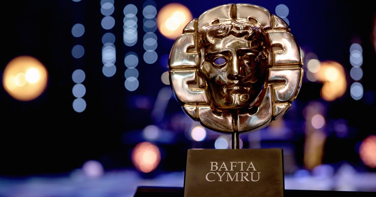A gold BAFTA Cymru mask trophy on a black plinth against a dark stage with blue and orange lights out of focus behind it