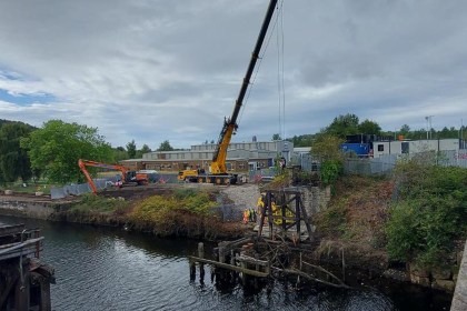 A crane and excavator on the bank of the River Tawe beside the Bascule Bridge, with workers in hi-vis alongside the old timber supports in the water