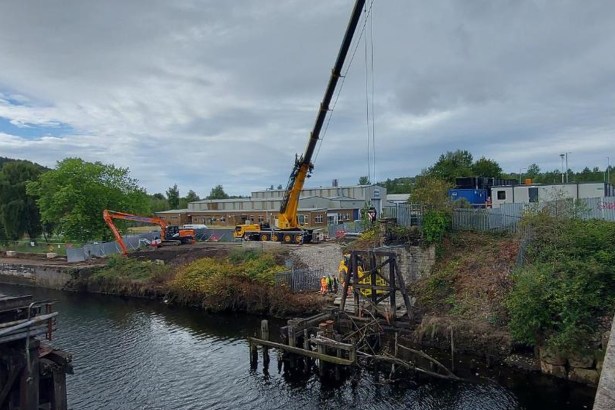 A crane and excavator on the bank of the River Tawe beside the Bascule Bridge, with workers in hi-vis alongside the old timber supports in the water