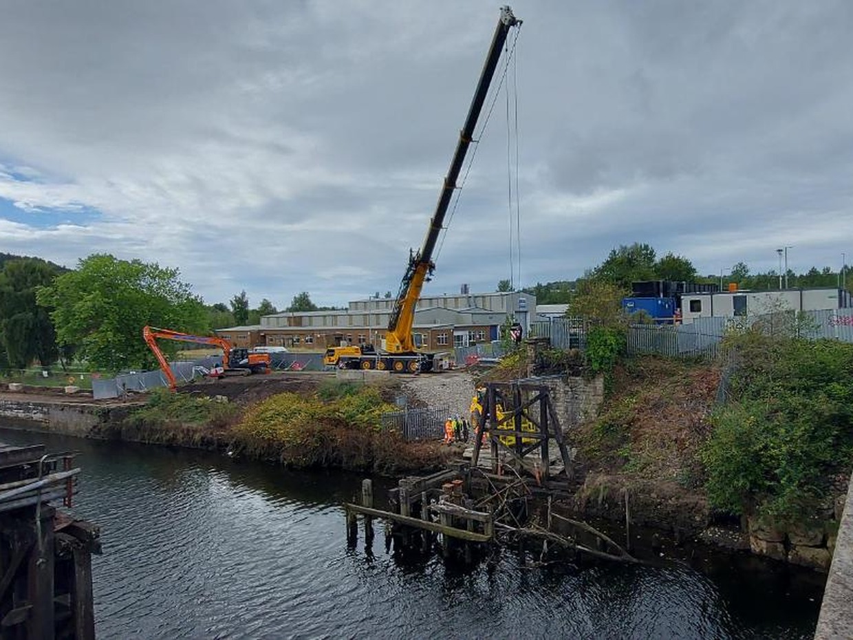A crane and excavator on the bank of the River Tawe beside the Bascule Bridge, with workers in hi-vis alongside the old timber supports in the water