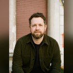 Author Ben Fergusson in a dark green corduroy jacket sitting in front of a red shutter
