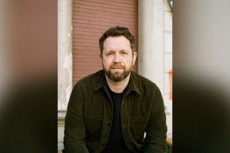 Author Ben Fergusson in a dark green corduroy jacket sitting in front of a red shutter