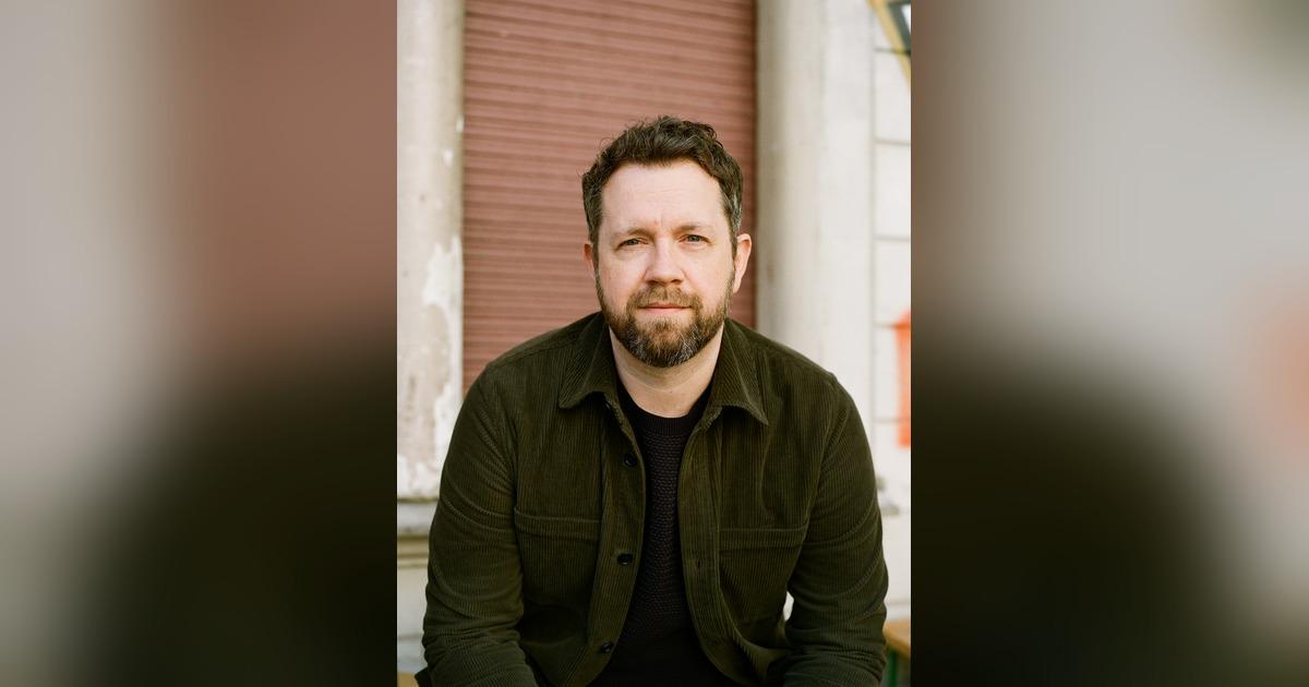 Author Ben Fergusson in a dark green corduroy jacket sitting in front of a red shutter