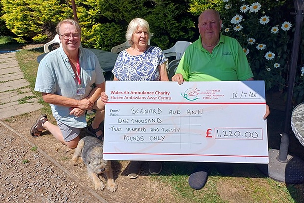 Ann and Bernard Williams kneeling in their garden with a large Wales Air Ambulance cheque for £1,220, with their son's rescue dog Kipper lying in front and fundraising manager Mike May alongside