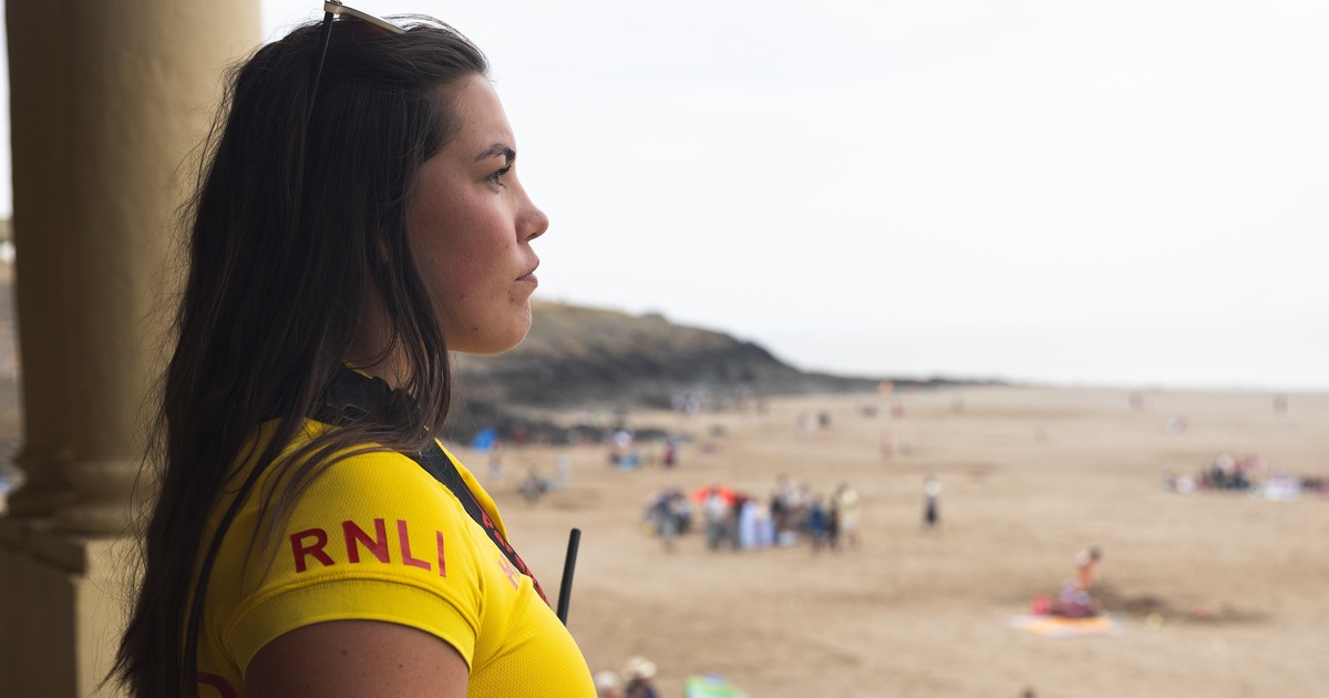 Beth Morgan in a yellow RNLI lifeguard top holding binoculars and looking out over a busy beach