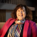 Bethan Evans smiling, seated in an armchair in a pink blazer and striped silk scarf