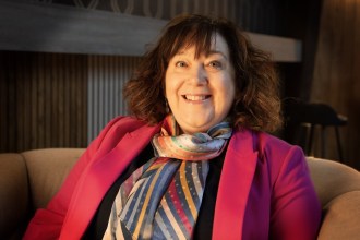 Bethan Evans smiling, seated in an armchair in a pink blazer and striped silk scarf