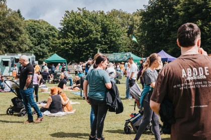 Crowds standing and sitting on grass at a street food festival, with food trucks and gazebos behind them and trees in the background