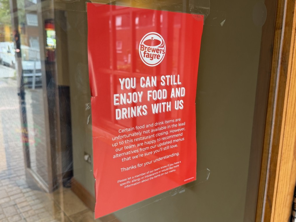 A red Brewers Fayre poster taped inside the Sandpiper's door reading you can still enjoy food and drinks with us, explaining some items are unavailable in the lead up to the restaurant closing