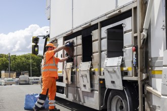 A worker in orange hi-vis clothing and a cap empties a recycling container into the side hatch of a kerbside recycling lorry, with blue recycling bags on the road beside him