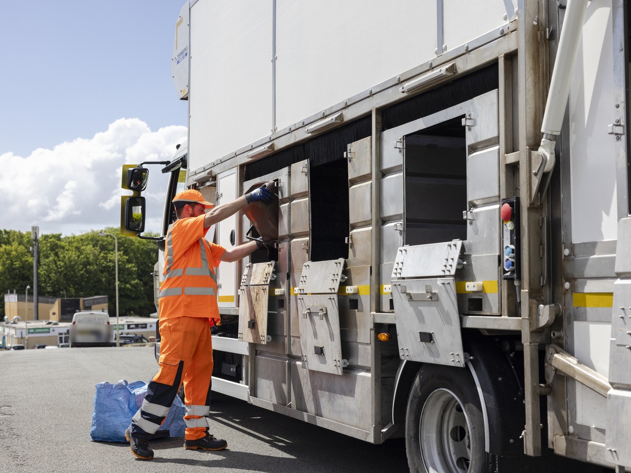 A worker in orange hi-vis clothing and a cap empties a recycling container into the side hatch of a kerbside recycling lorry, with blue recycling bags on the road beside him