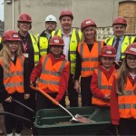 Four primary pupils in red hard hats and hi-vis vests with a spade and wheelbarrow, standing with adults in hard hats on a school building site