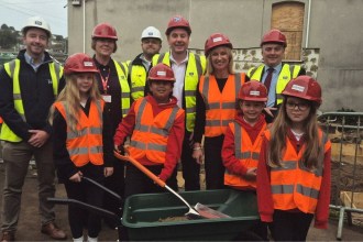 Four primary pupils in red hard hats and hi-vis vests with a spade and wheelbarrow, standing with adults in hard hats on a school building site