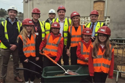 Four primary pupils in red hard hats and hi-vis vests with a spade and wheelbarrow, standing with adults in hard hats on a school building site