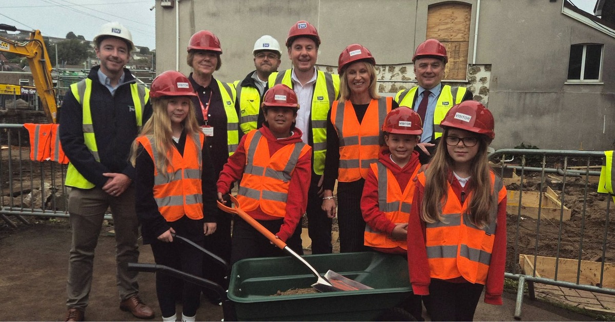 Four primary pupils in red hard hats and hi-vis vests with a spade and wheelbarrow, standing with adults in hard hats on a school building site