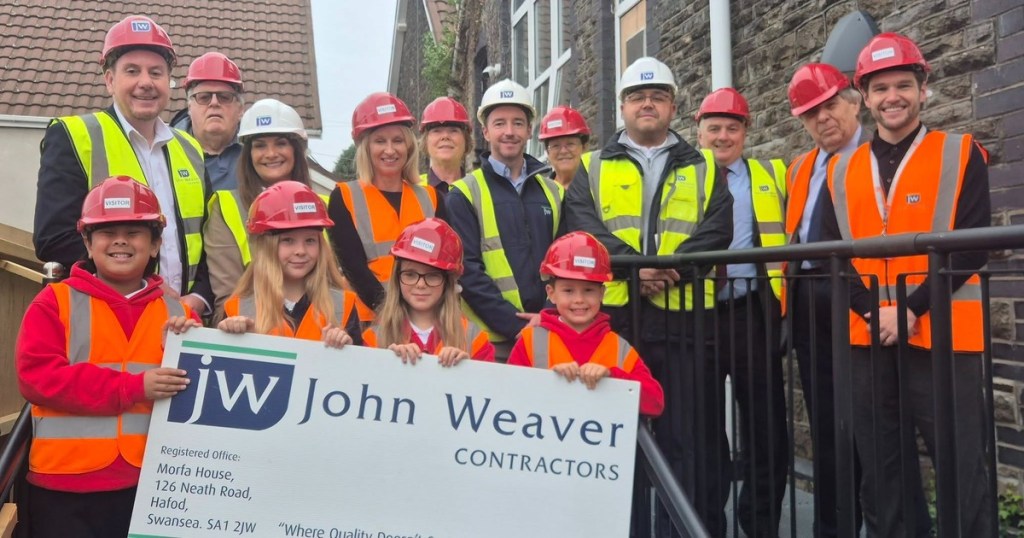 Four primary pupils in hard hats holding a John Weaver Contractors sign, with a group of adults in hi-vis behind them outside a stone school building