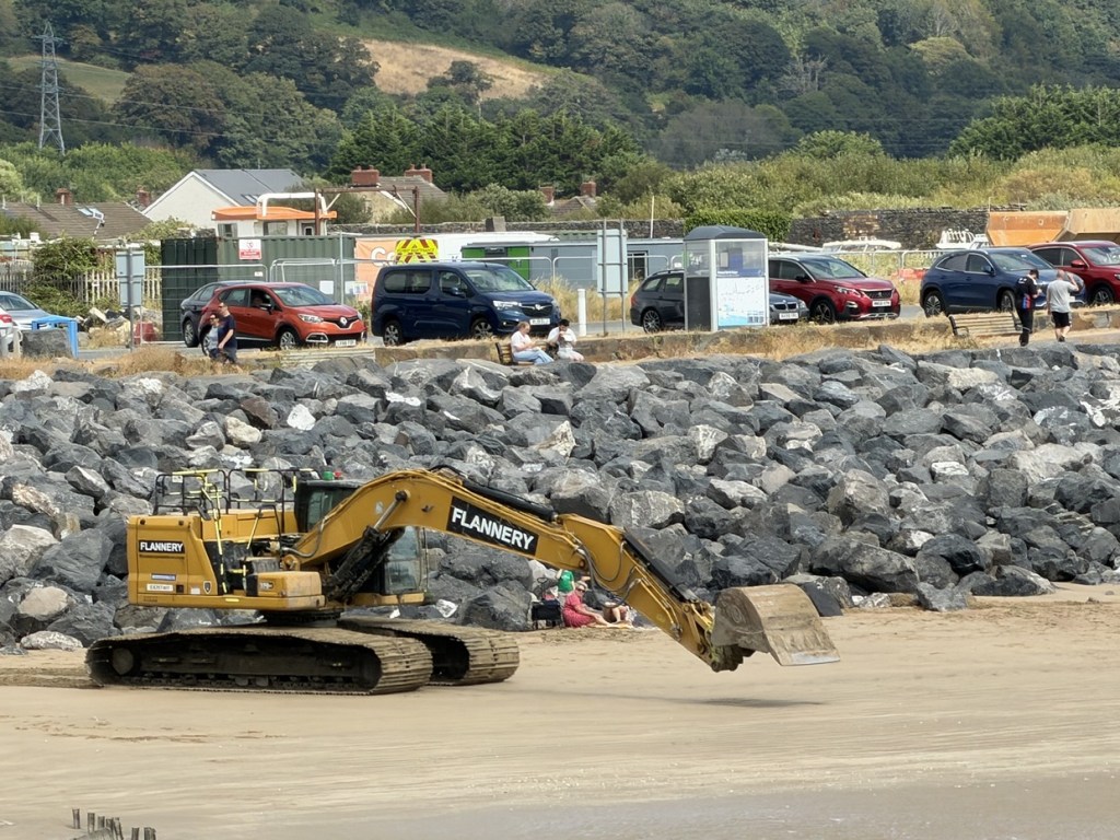 A Flannery excavator working on the sand below the rock armour at Burry Port, with people sitting on benches behind