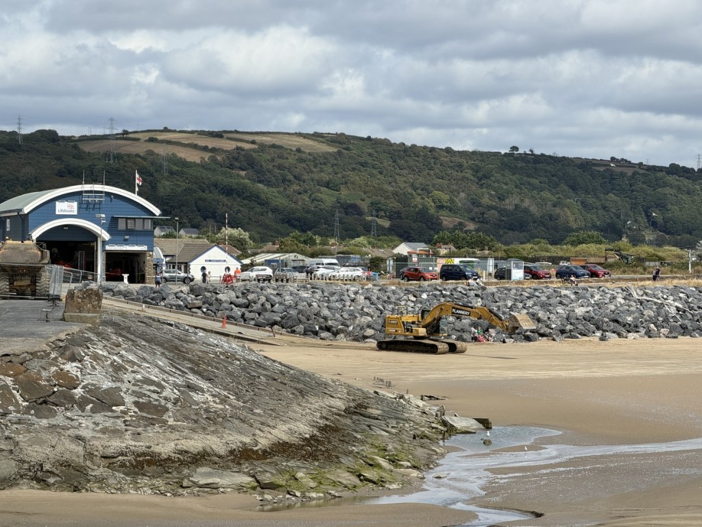 An excavator on the beach beside Burry Port's lifeboat station with the hillside behind