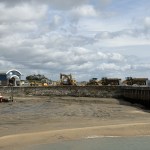 An excavator and dump trucks lined along the quay at Burry Port Harbour, with a red fishing boat aground on the drained harbour bed below