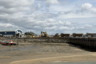 An excavator and dump trucks lined along the quay at Burry Port Harbour, with a red fishing boat aground on the drained harbour bed below