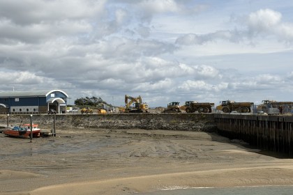 An excavator and dump trucks lined along the quay at Burry Port Harbour, with a red fishing boat aground on the drained harbour bed below