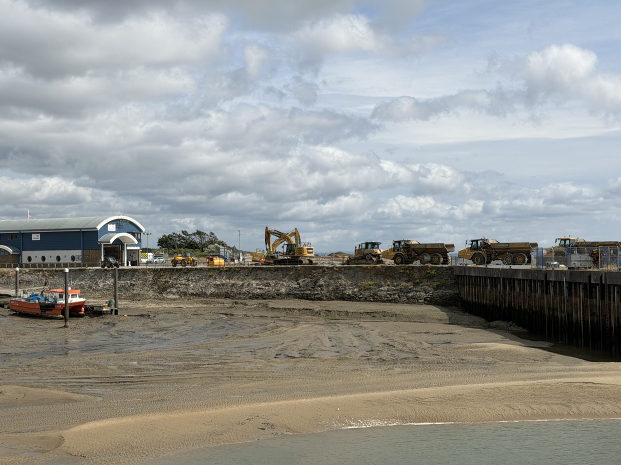 An excavator and dump trucks lined along the quay at Burry Port Harbour, with a red fishing boat aground on the drained harbour bed below