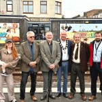 Ten people, including a mayor in chains and a woman holding a toddler, lined up on a railway platform in front of two colourful Welcome to Burry Port and Welcome to Pembrey murals