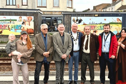 Ten people, including a mayor in chains and a woman holding a toddler, lined up on a railway platform in front of two colourful Welcome to Burry Port and Welcome to Pembrey murals