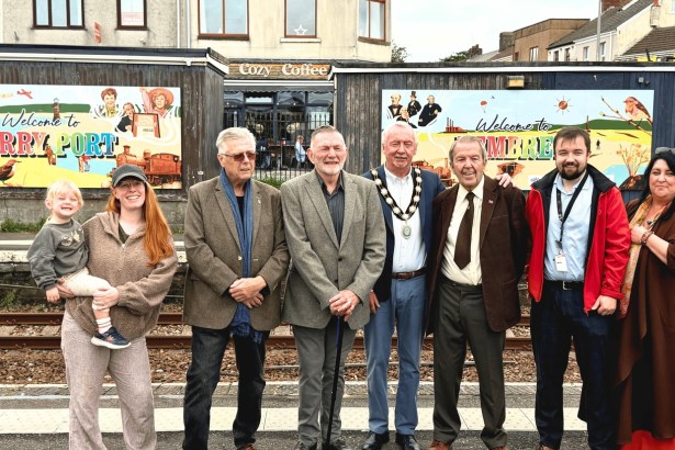 Ten people, including a mayor in chains and a woman holding a toddler, lined up on a railway platform in front of two colourful Welcome to Burry Port and Welcome to Pembrey murals