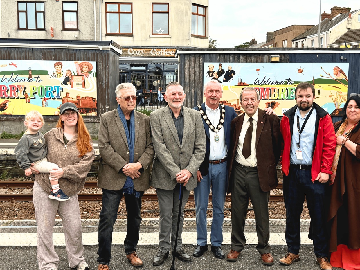 Ten people, including a mayor in chains and a woman holding a toddler, lined up on a railway platform in front of two colourful Welcome to Burry Port and Welcome to Pembrey murals