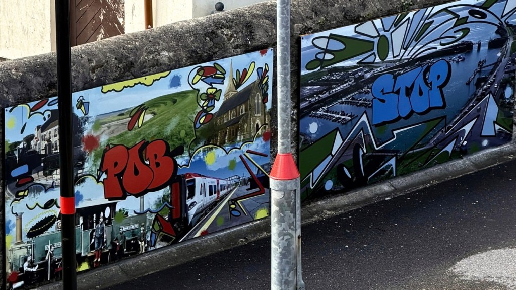 Two graffiti-style panels on a stone wall reading Pob and Stop, with photographs of a church, a train, a steam locomotive crew and the harbour worked into the design