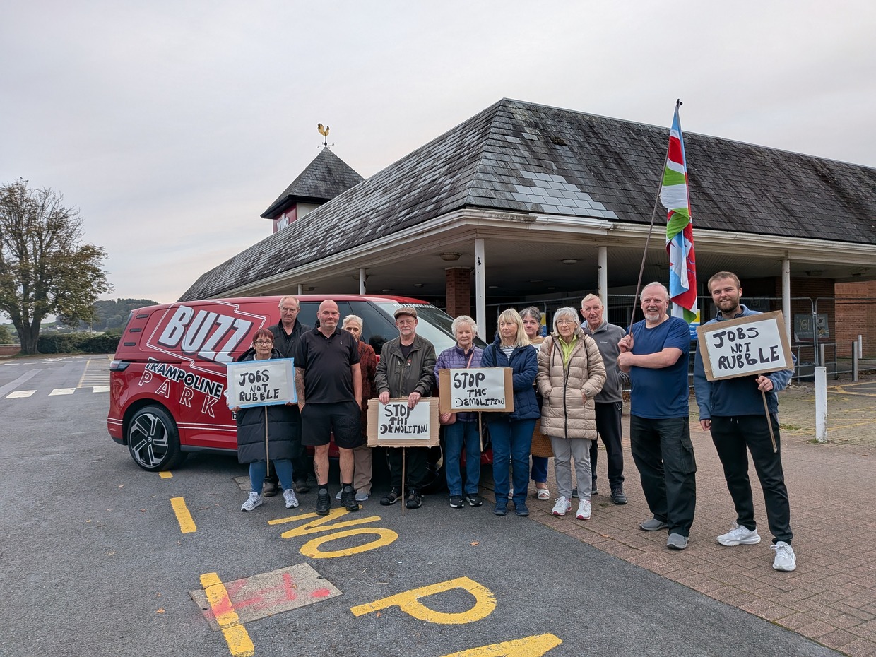 A group of campaigners holding "Jobs not rubble" and "Stop the demolition" placards beside a red Buzz Trampoline Park van outside the former Wilko store in Carmarthen