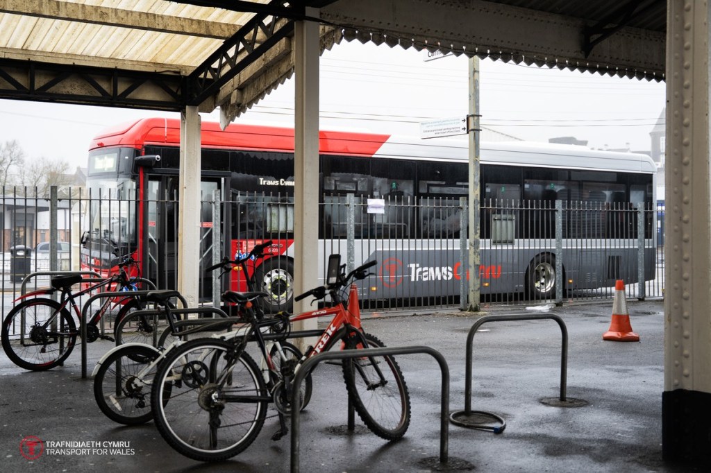Bicycles locked to hoops under Carmarthen station's canopy, with a red and grey TrawsCymru bus visible through the railings outside