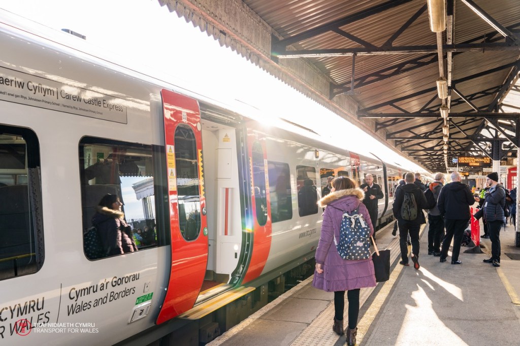 Passengers walking along a covered platform at Carmarthen railway station beside a white and red Transport for Wales train with its doors open