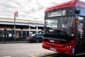 A red TrawsCymru T1 bus signed Lampeter for Aberystwyth pulling away from the kerb outside Carmarthen railway station, with the platform canopy and taxi rank behind