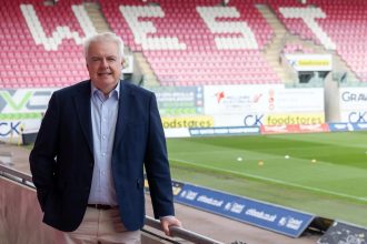 Former First Minister Carwyn Jones in a navy blazer pitchside at Parc y Scarlets, with rows of red seats behind him